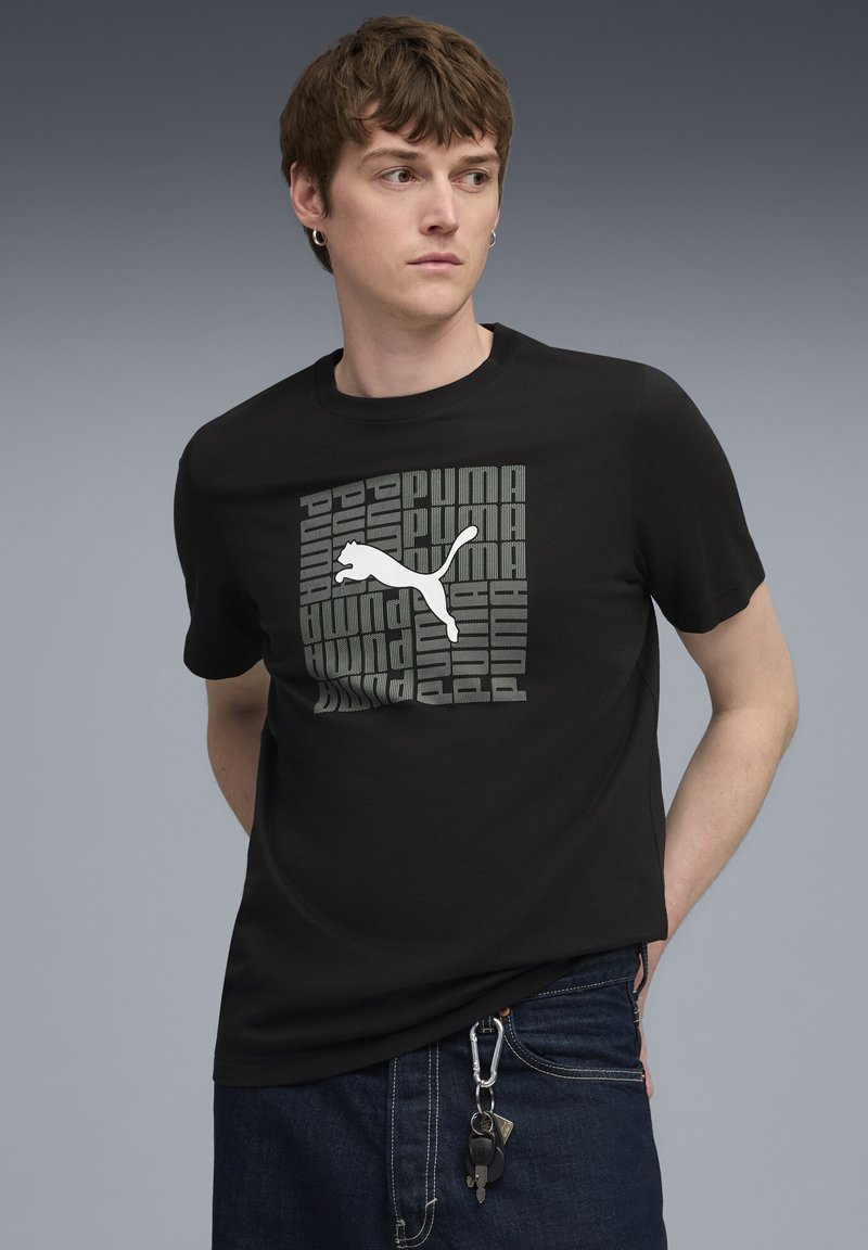 Camiseta de algodón negra con un gran logo plateado de Puma y un diseño de texto repetido. Cuello redondo y mangas cortas. Llave visible en los vaqueros.