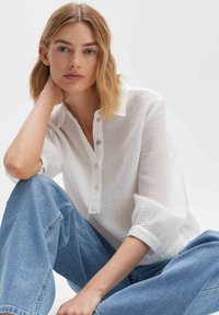 Camicia bianca a maniche lunghe con texture e colletto, caratterizzata da bottoni pastello. Abbinata a jeans in denim blu chiaro a vita alta, in posizione seduta.