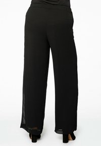 Zwarte wijde broek van lichtgewicht, gestructureerd materiaal met een elastische tailleband. Heeft een doorschijnende overlay langs de zijkanten.