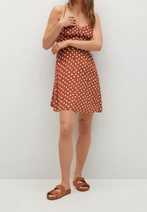 Ruskea hihaton mekko, jossa on valkoisia pilkkuja, etuosassa solmittu yksityiskohta ja levenevä helma. Yhdistetty ruskeisiin sandaaleihin. Sileä kangastuntu.