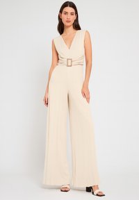 Jumpsuit beige con scollatura a V profonda, design plissettato e una fibbia decorativa in vita. Le gambe larghe si estendono per coprire le scarpe.