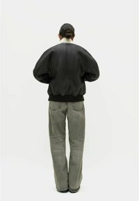 Schwarze Lederbomberjacke mit einem cremefarbenen Kragen, oversize Schnitt und gerippten Bündchen. Graue, locker sitzende Jeans mit sichtbaren Nähdetails.