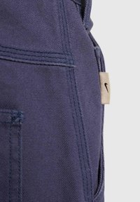 Nike kombinezoni iz temno mornarsko modrega platna, z vidnim šivanjem, stranskim žepom in majhno bež logotipno etiketo.