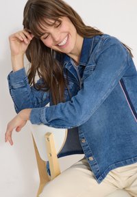 Jeune femme en veste en jean assise de côté sur une chaise en bois, souriant les yeux fermés devant un fond uni.