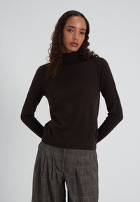 Femme aux cheveux bouclés portant un pull à col roulé marron foncé et un pantalon large à carreaux gris, se tenant avec les mains derrière le dos.