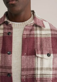 Rotes und cremefarbenes kariertes Hemd mit Fischgratmuster, schwarzen Knöpfen und einem beigen Strickpullover darunter mit einem gerippten Kragen.