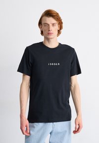 Juodas medvilninis T-shirt su trumpomis rankovėmis ir apvalia iškirpte, ant krūtinės esantis žodis "JORDAN" parašytas ryškiu, baltu, stilingu šriftu.