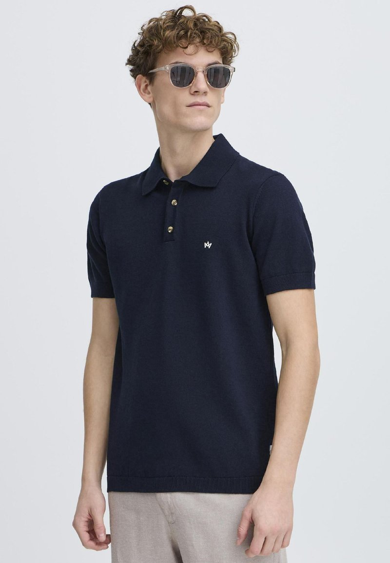 Polo de culoare bleumarin, din material tricotat, cu guler clasic, închidere cu trei nasturi și un logo mic pe piept. Mâneci scurte.