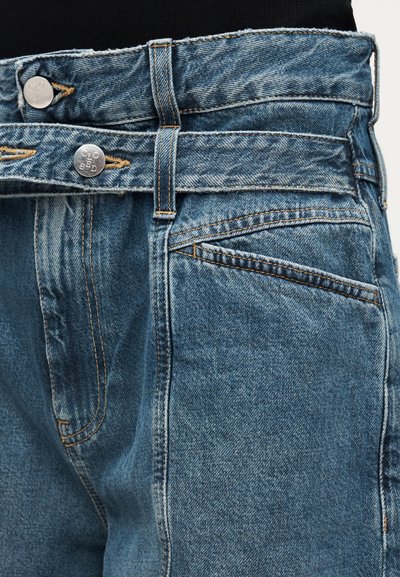 Κοντινή λήψη διπλής μέσης μπλε τζιν τζιν με ασημένια κουμπιά και πορτοκαλί ραφή σε μαύρο ύφασμα.