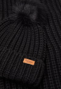 Caciulă din tricot neagră cu un pompon din blană, având un model texturat cu nervuri. O etichetă din piele bej în partea de jos spune "Barbour England."