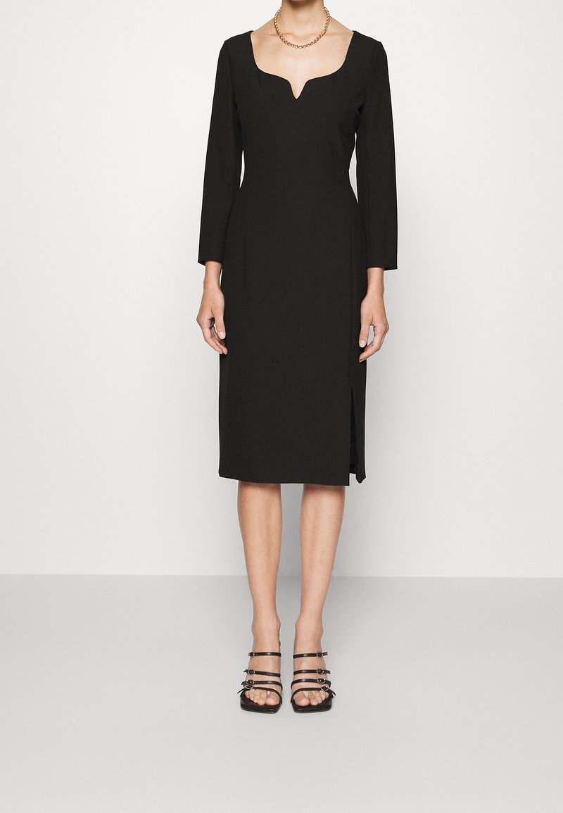 Femme portant une robe midi noire ajustée avec manches longues, un décolleté en forme de cœur, une fente latérale et des sandales à talons noires avec lanières.