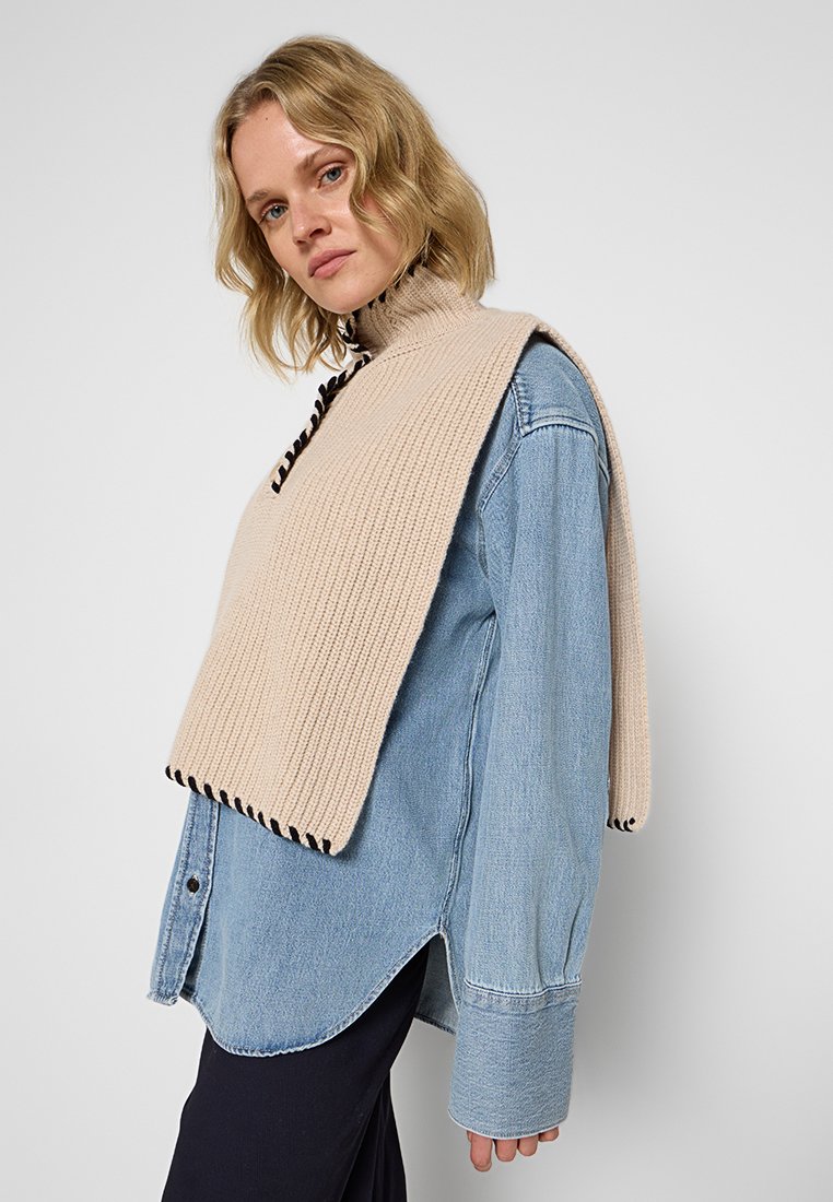 Beige gestrickter Schal mit schwarzen Akzenten über einem hellblauen Denimhemd. Der Schal hat eine strukturierte Oberfläche und ein asymmetrisches Design.