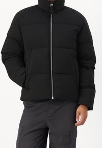 Osoba ubrana w czarną pikowaną kurtkę z przednim suwakiem i ciemne spodnie bojówki, ręce luźno opuszczone wzdłuż ciała.