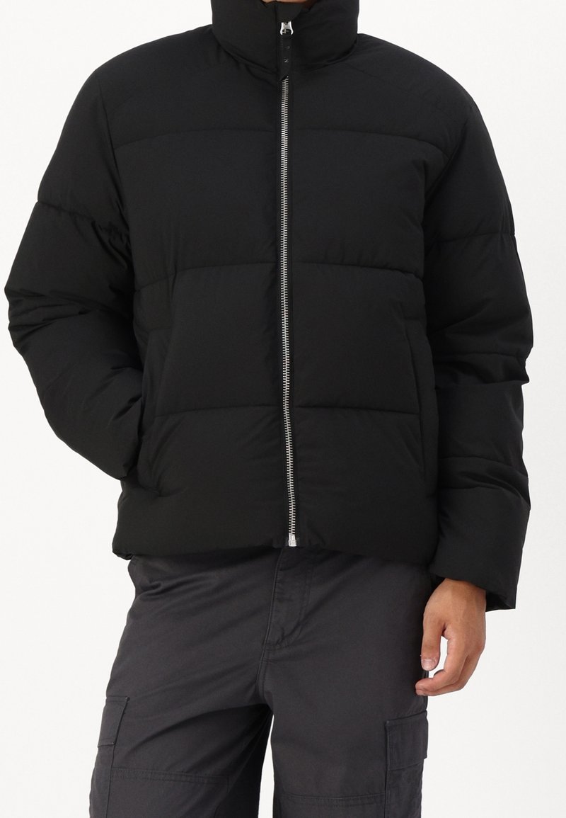 Osoba ubrana w czarną pikowaną kurtkę z przednim suwakiem i ciemne spodnie bojówki, ręce luźno opuszczone wzdłuż ciała.