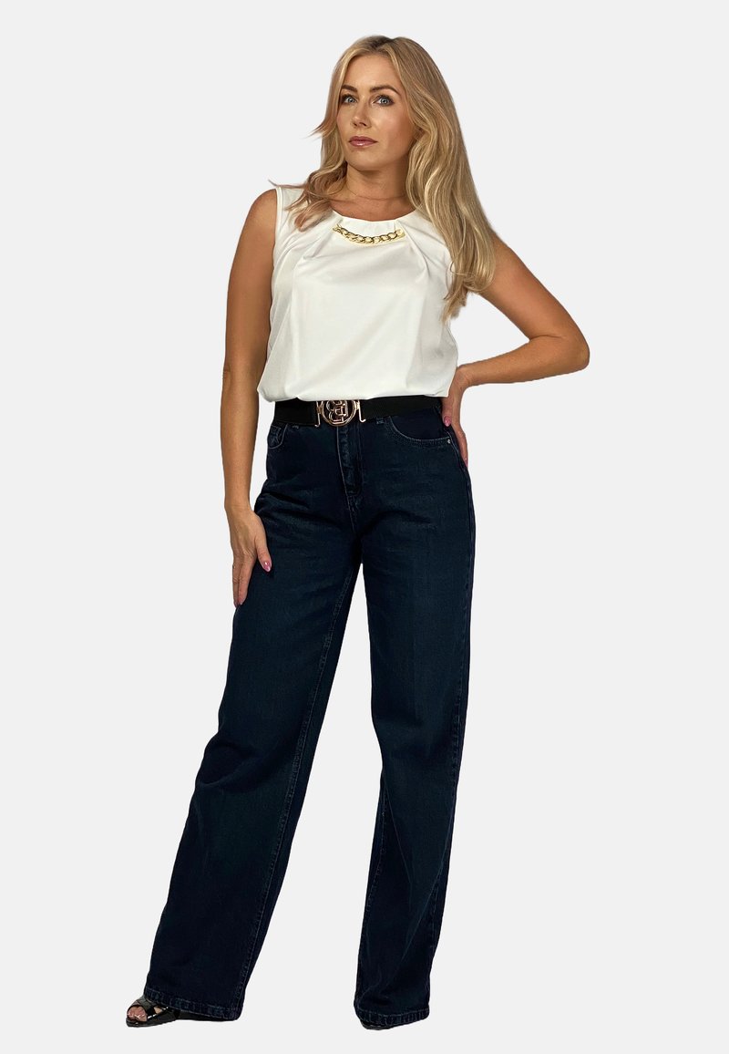 Biała bezrękawnik z detalem w postaci złotego łańcuszka, w połączeniu z ciemnoniebieskimi spodniami o szerokiej nogawce. Modelka nosi czarny pasek z złotą klamrą.