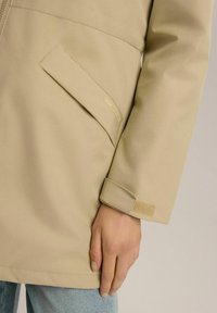 Beżowa, wodoodporna kurtka z zapięciem na zamek, z boczną kieszenią, rzepem przy mankietach i gładką, miękką fakturą.