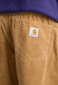 Fløjlsbukser i beige med fremtrædende vertikale riller, der har et hvidt mærke med en orange logo på baglommen. Synlig sømning.