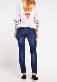Suéter de punto gris claro con un profundo escote en V en la espalda y detalle de tirantes, combinado con unos jeans ajustados de color azul oscuro. El suéter tiene mangas largas y acanaladas.