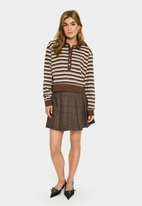 Femme debout portant un polo à manches longues rayé marron et beige, une jupe plissée à carreaux marron et des escarpins à bout pointu noirs.