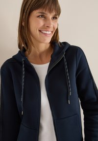 Donna sorridente con capelli castani che indossa una felpa con zip blu navy sopra una maglietta bianca, su uno sfondo chiaro e semplice.