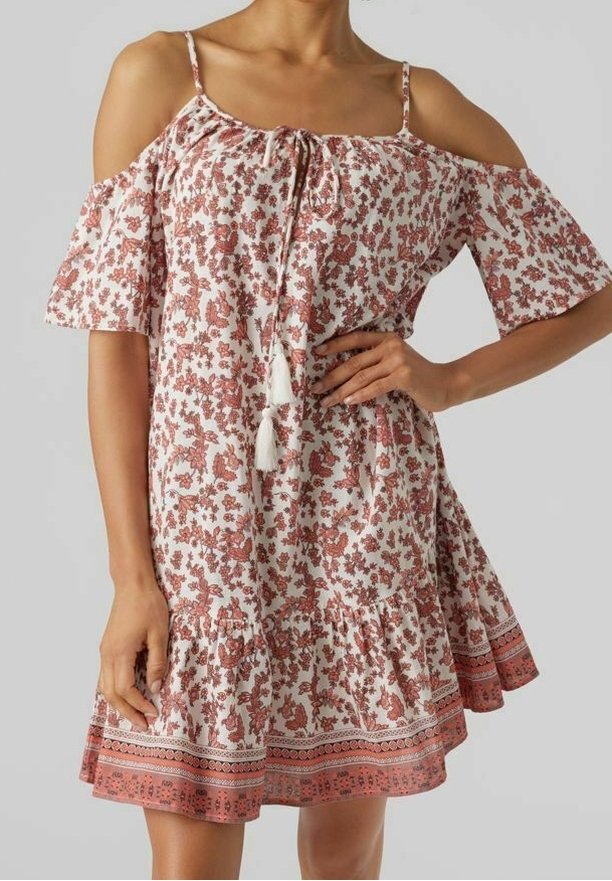 Femme portant une robe blanche et rouille à fleurs, épaules dénudées, avec bretelles spaghetti et liens à pompons au niveau de l'encolure.