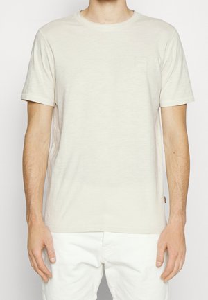 Mann trägt ein schlichtes, hellbeiges T-Shirt mit kurzen Ärmeln und weiße Hose, steht mit entspannten Armen seitlich an einem hellen Hintergrund.