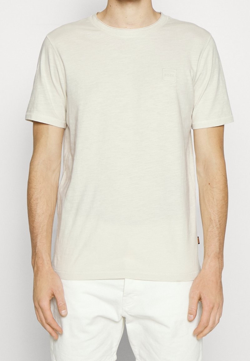 Homme portant un t-shirt uni beige clair à manches courtes et un pantalon blanc, debout, les bras détendus le long du corps, sur un fond clair.