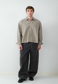 Karierte beige Langarmbluse mit Tasche, kombiniert mit locker sitzenden dunkelgrauen Jeans. Schwarze Schuhe runden das Outfit ab.