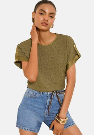 Kobieta ubrana w oliwkowy, teksturowany top z krótkim rękawem, z mankietami podwiniętymi i zapinanymi na guziki oraz niebieskie, wysokiego stanu szorty jeansowe z wzorzystym paskiem.