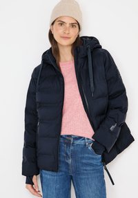 Marineblå pufferjakke med hætte, lynlås, elastiske manchetter og et logo på ærmet. Bæres over en lyserød sweater og blå jeans.