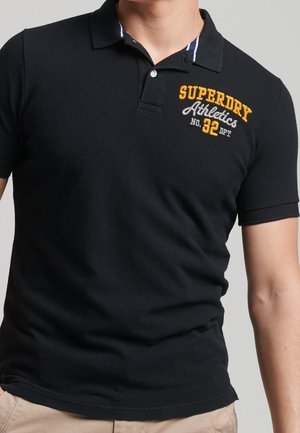 Mężczyzna ubrany w czarną koszulkę polo z haftowanym na piersi żółto-białym napisem "Superdry Athletics No. 32 DPT".