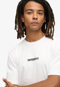 Hvid bomulds t-shirt med korte ærmer, med sort "SUPERDRY®" logo broderi på brystet. Glat tekstur, standard pasform.