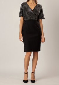 Femme portant une robe noire jusqu'aux genoux avec un haut texturé métallique en style wrap et des manches bouffantes, associée à des talons à bride chevillère noirs.