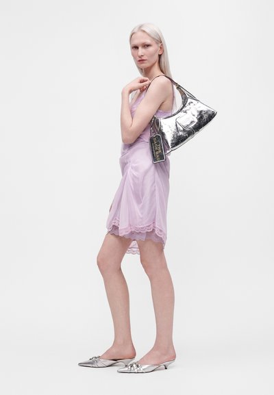 Femme en robe nuisette lilas bordée de dentelle, portant des chaussures pointues argentées et un sac à main argenté métallique sur l'épaule, sur un fond uni.