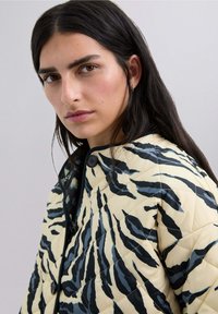 Chaqueta acolchada en crema con rayas de tigre en negro y azul, con un cuello redondo, dos botones y un acabado suave y brillante.