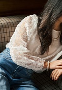 Haut blanc avec manches bouffantes en dentelle transparente ornées de broderies florales délicates, associé à un jean en denim bleu et accessoirisé d'un bracelet en perles.