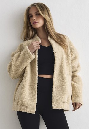 Kobieta o długich blond włosach, ubrana w beżowy płaszcz typu teddy na czarnym krótkim topie i czarnych spódnicach z wysokim stanem, stoi na tle białej ściany.