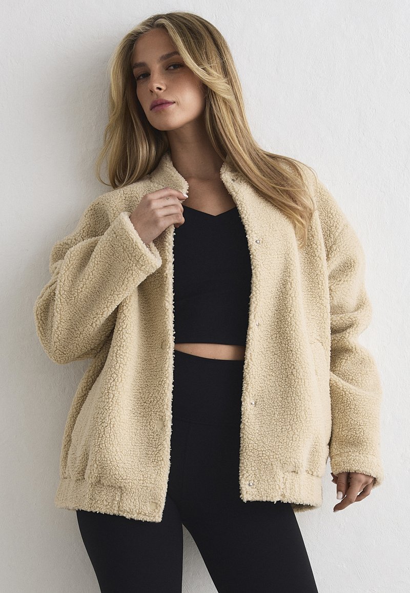 Naine pika blondi juustega, kes kannab beežikat teddy-mantlit musta lühikese toppi ja kõrge vöökohaga mustade pükste kohal, seistes valge seina taustal.