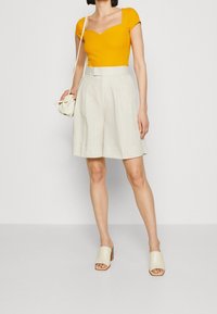 Gul ribbad kortärmad topp med fyrkantig halsringning, tillsammans med beige linshorts med ett bälte. Ljusfärgade block-klack-mules bärs.