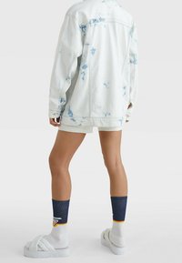 Weiße Jeansjacke mit blauen Batikmustern, kombiniert mit hellen Shorts, weißen Slide-Sandalen und marineblauen Socken mit orangen Akzenten.