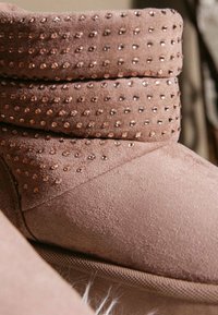 Stivaletto alla caviglia in pelle scamosciata rosa con risvolto decorato da borchie color oro rosa. Lo stivale ha una texture morbida e una forma con punta arrotondata.