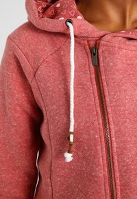 Sudadera con capucha de forro polar rojo con cierre de cremallera, cordones blancos y un forro interior estampado con pequeños corazones. Tejido suave y texturizado.