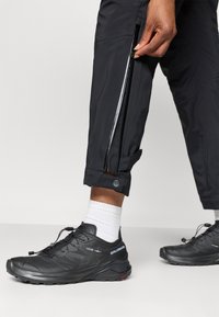 Pantalones impermeables negros con bolsillos laterales con cremallera y puños con botón, combinados con zapatillas deportivas negras que cuentan con un cierre de cordones y suela texturizada.