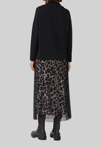 Femme vêtue d'un haut noir à manches longues, d'une jupe mi-longue plissée à imprimé léopard et de bottines noires, de dos devant un fond uni.