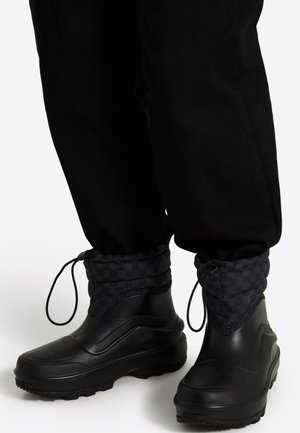 Black waterproof ankle boots with textured soles and adjustable drawstring cuffs worn with black pants.