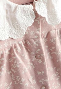 Růžové šaty z látky s bílým květinovým vzorem; mají bílý krajkový límeček a detail knoflíků na přední straně. Měkká textura, nabraný pas.