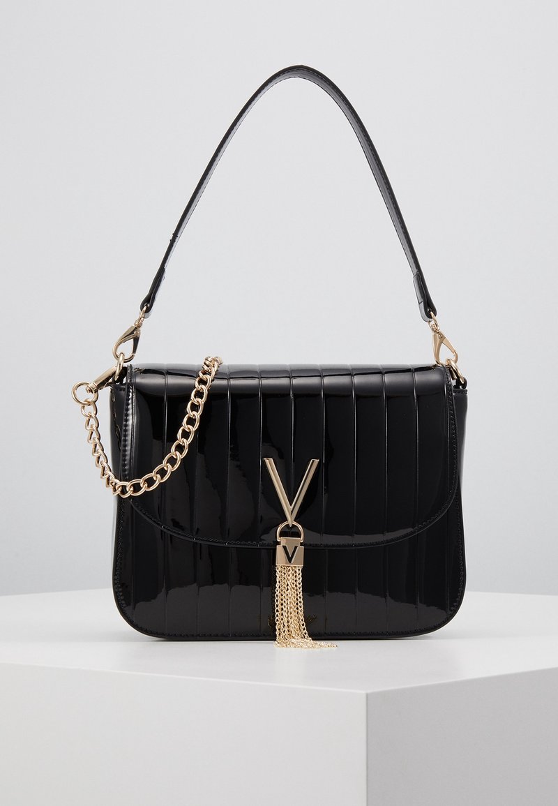 Black glossy handbag with vertical stitching, gold chain strap, and V-shaped gold clasp with tassel, placed on white surface.