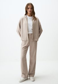 Beige fleece zip-up hoodie en bijpassende joggerbroek met trekkoord, met een ontspannen fit en een gladde textuur, gecombineerd met witte sneakers.