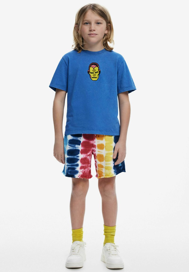 Blue cotton t-shirt with a yellow character print on the chest, paired with multicolored tie-dye shorts. White sneakers and yellow socks.
