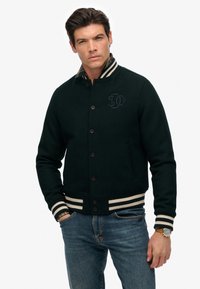 Mørkegrøn bomberjakke med krave, prydet med et logo på forsiden, sorte knapper og stribede strikkede manchetter og kant. Båret med blå jeans.